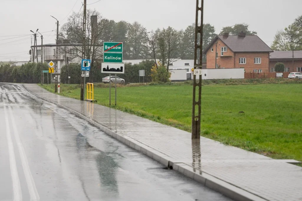 Deszczowy dzień na skraju polskiej miejscowości: mokra droga, chodnik, tablice kierunkowe na Będków i Kalinów oraz zabudowania jednorodzinne.