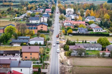Widok z lotu ptaka na wieś rozciętą główną drogą, po której jeżdżą samochody, z robotami drogowymi po jednej ze stron i zabudową jednorodzinną w otoczeniu pól o