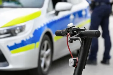 Kierownica hulajnogi elektrycznej na pierwszym planie, w tle policyjny radiowóz i stojący funkcjonariusz.