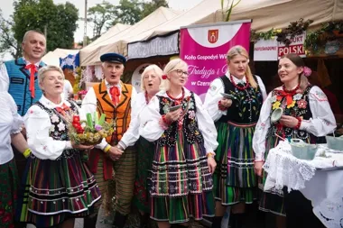 Grupa osób w strojach ludowych stoi przed namiotami z regionalnymi wyrobami podczas plenerowej imprezy kulturalno-gastronomicznej.