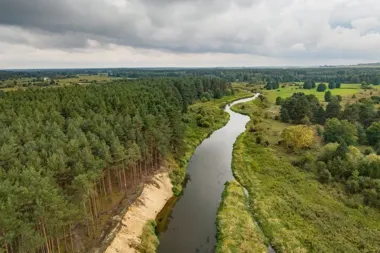 Krzewiasta rzeka płynie przez zalesioną i zieloną dolinę pod zachmurzonym niebem.