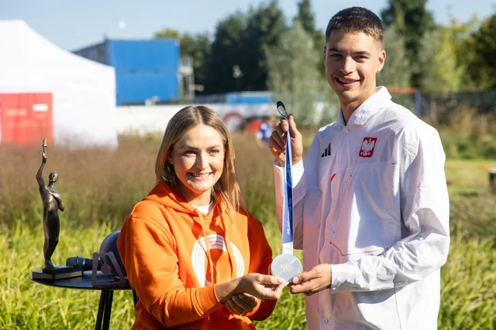 Kobieta w pomarańczowej bluzie i mężczyzna w białej kurtce z polskim godłem trzymają srebrny medal na niebieskim pasku, stojąc na trawie na tle namiotów i zabud
